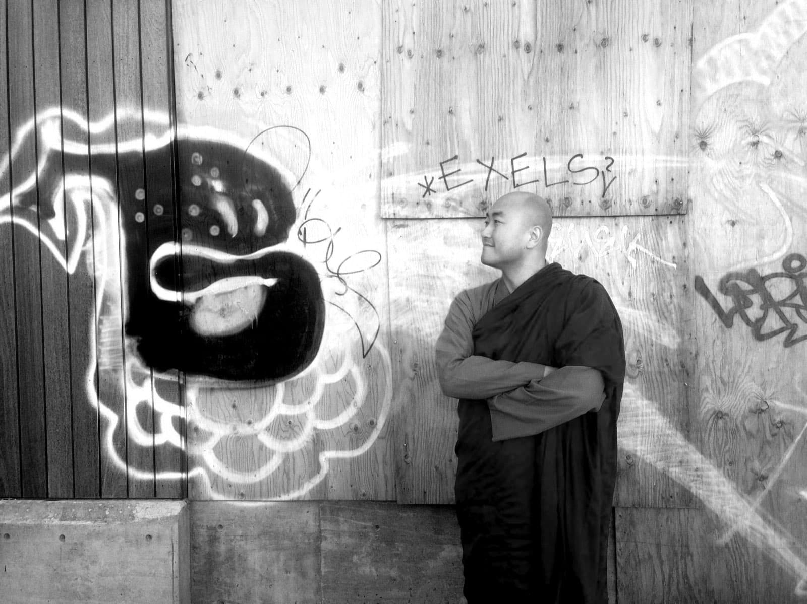 Sagarananda Tien standing before a graffitied plywood wall
